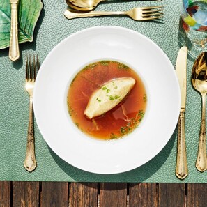 A white plate with a soup and a piece of meat on a green textured tablecloth.