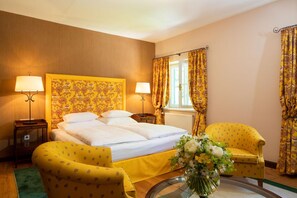 A hotel room with a large bed, two bedside lamps, a floral-patterned headboard, a yellow armchair, a round coffee table, and a vase with flowers.