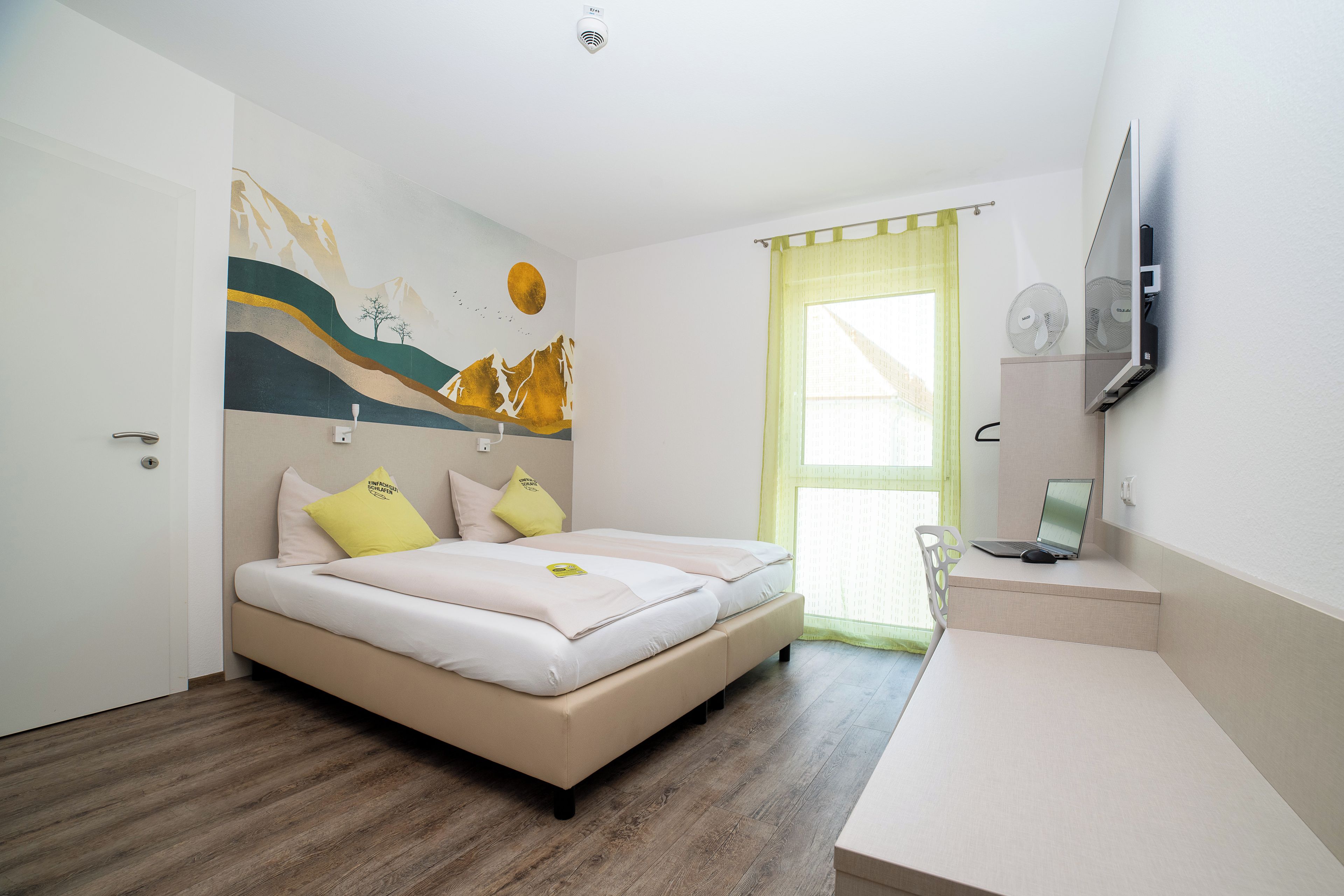 Un dormitorio con cama, escritorio, computadora portátil, televisor y ventana con cortinas.