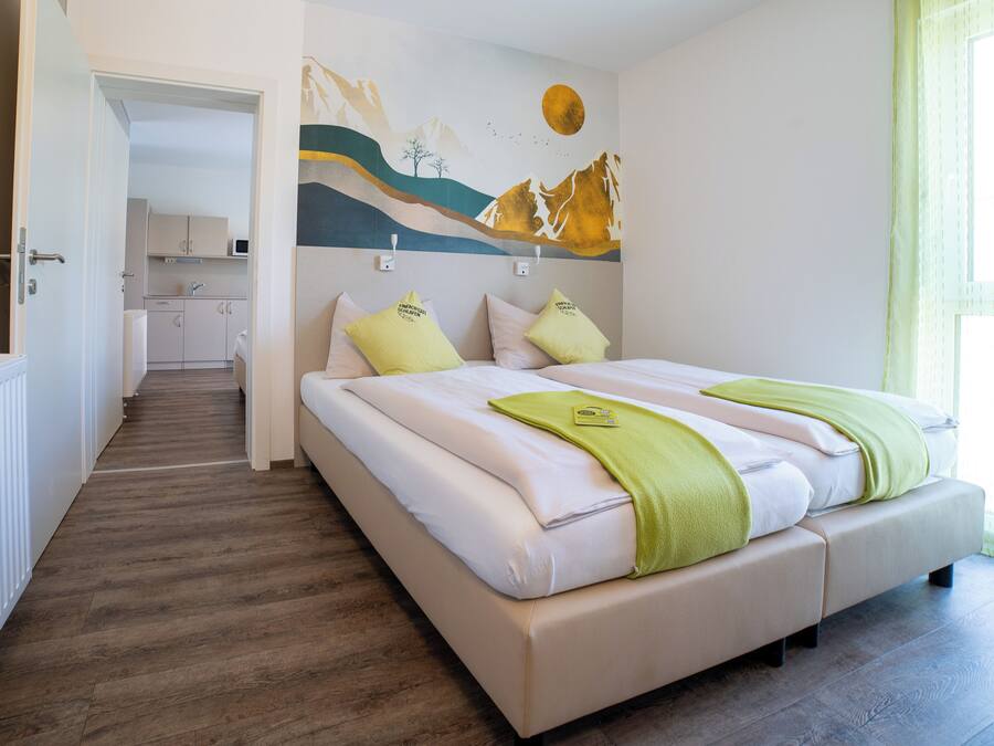 A bedroom with a bed, pillows, a green towel, a wall mural, and a view of a kitchen.