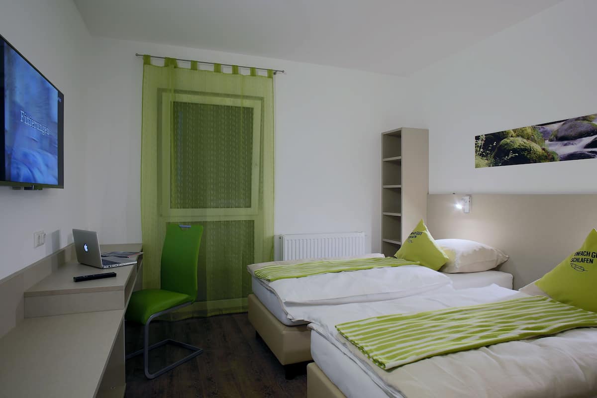 Habitación de hotel con dos camas, un escritorio con una laptop, una silla verde, un televisor y una ventana con cortina.