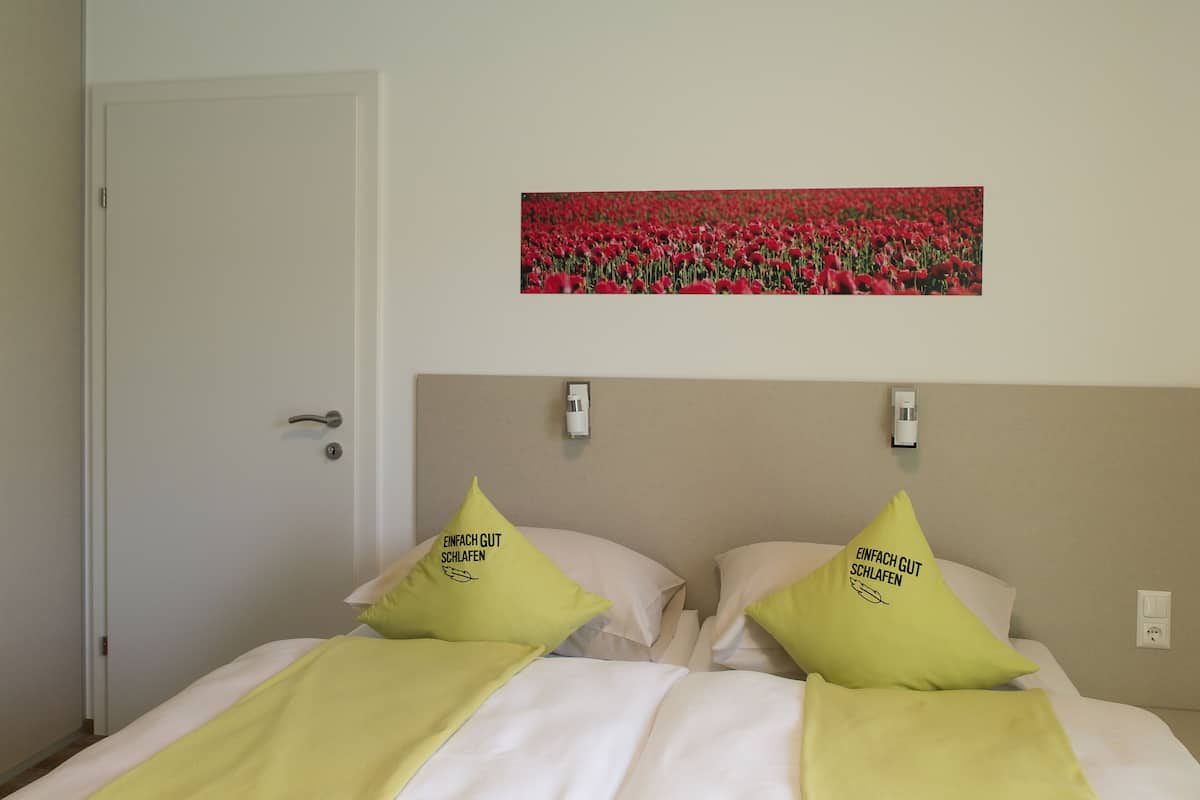 Un dormitorio con una cama, dos almohadas con texto, un cuadro de flores rojas colgado en la pared y una puerta.