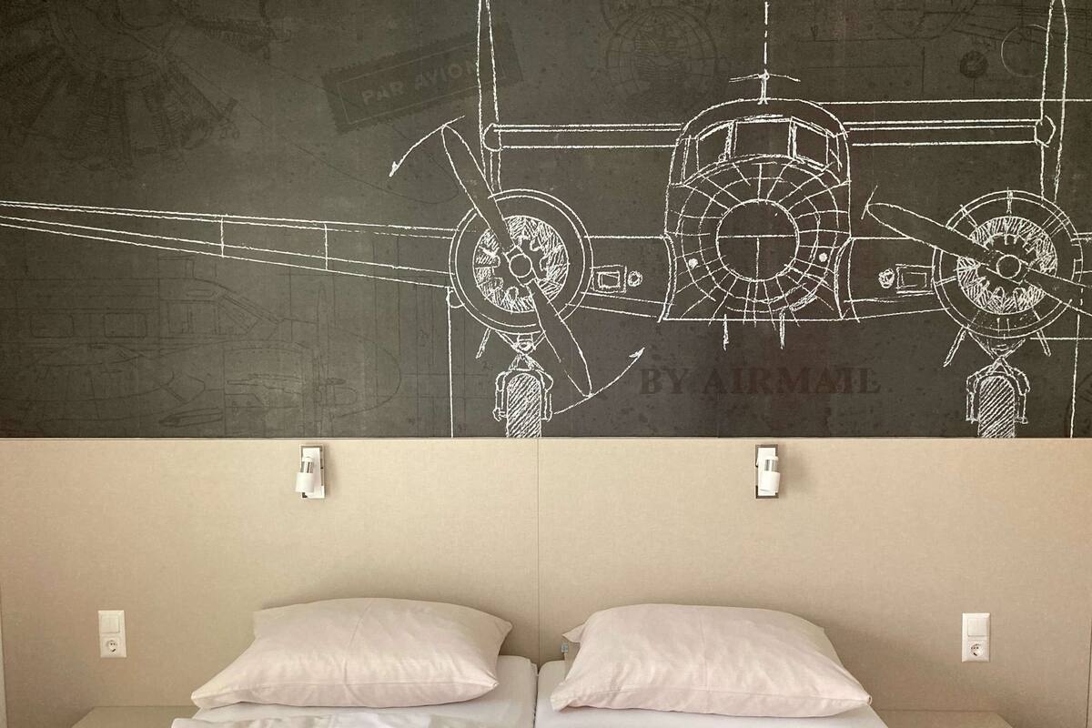 Habitación de hotel con una cama, dos mesitas de noche y una pared de pizarra con dibujos técnicos.