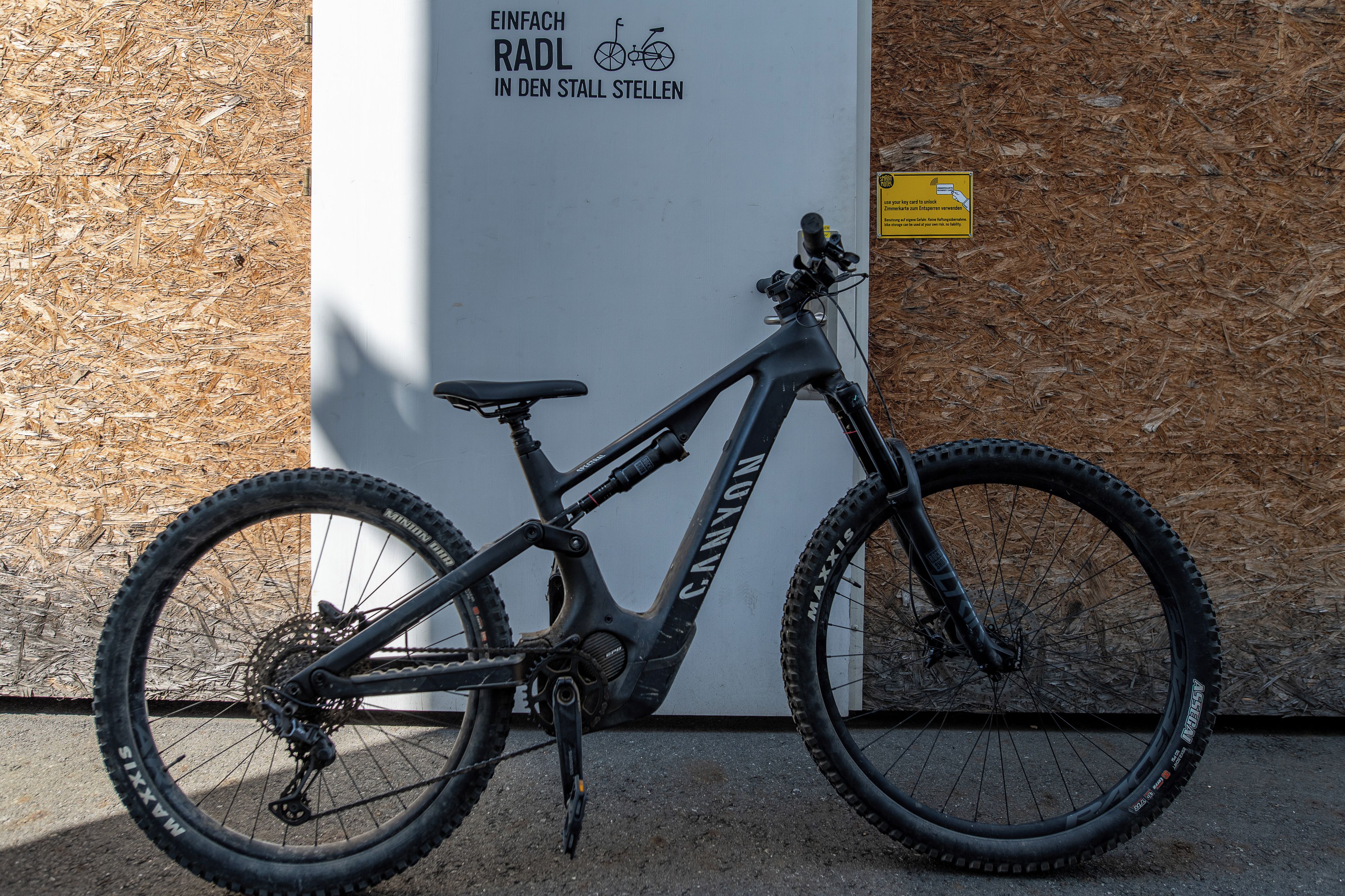 A mountain bike parked against a wall with a sign that reads 'Einfach RADL IN DEN STALL STELLEN'.