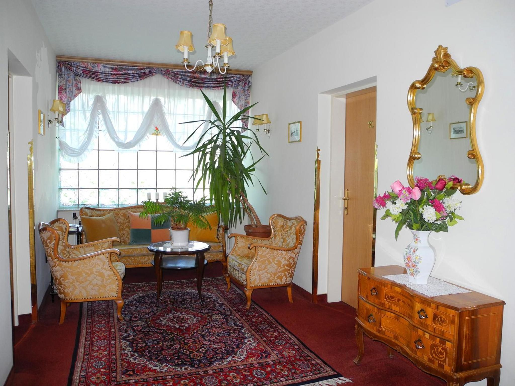 Ein Zimmer mit einer Couch, zwei Sesseln, einem Couchtisch, einer großen Topfpflanze, einem Spiegel und einer Kommode mit Blumen.