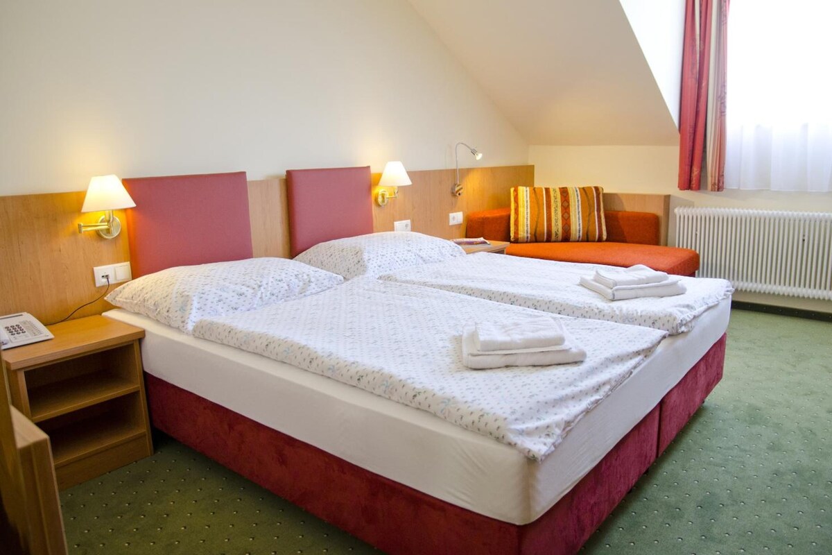 A hotel room with two beds, a red headboard, a small bedside table with a lamp, and a window with curtains.