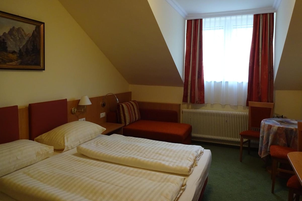 A hotel room with a pitched ceiling, a bed with two pillows, a bedside table with a lamp, a window with curtains, and a radiator.