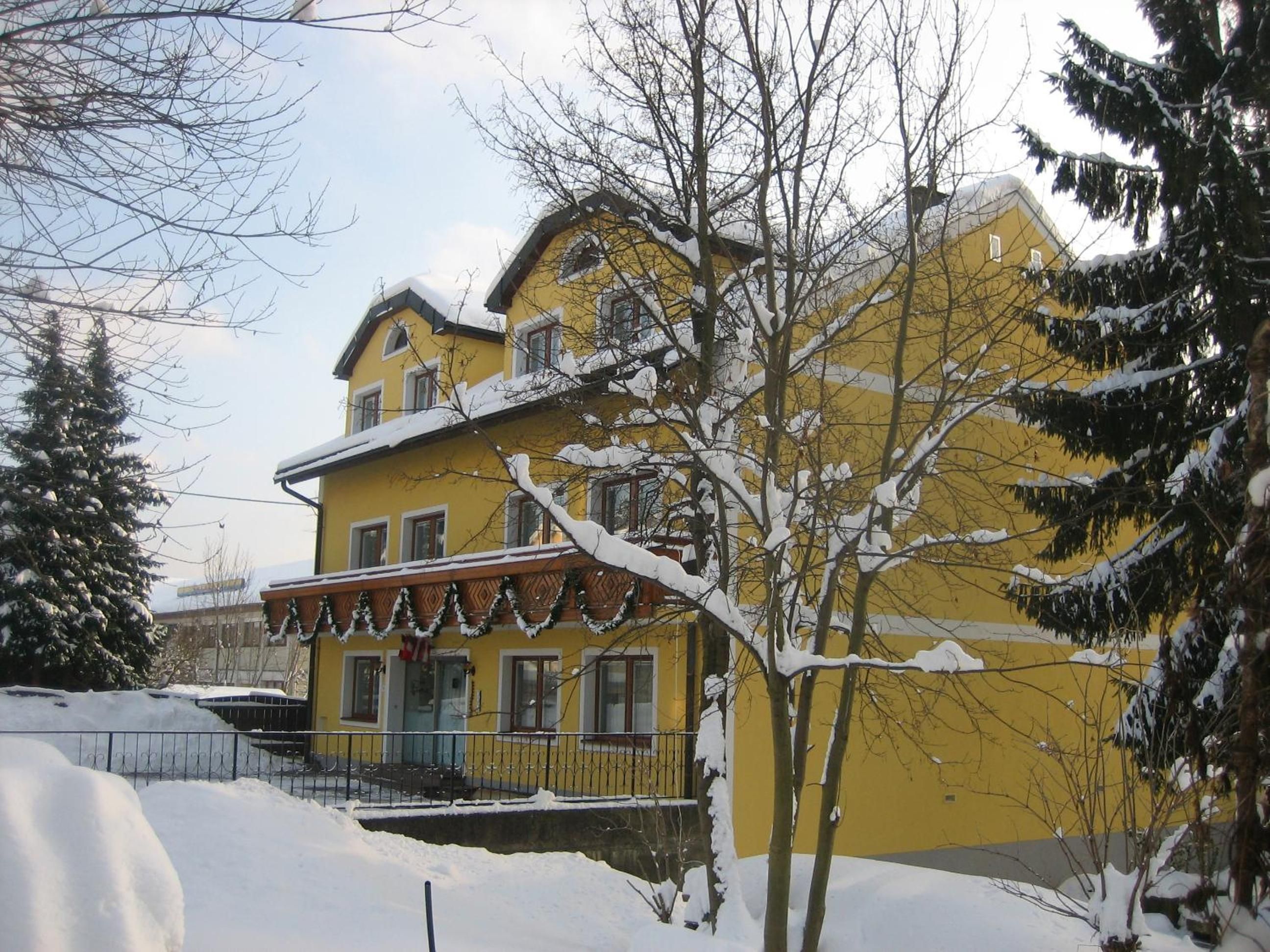 Ein zweistöckiges Gebäude mit einer gelben Fassade, einem Balkon mit Geländer und Schnee auf dem Dach und den umliegenden Bäumen.