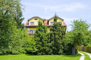 A two-story house with a yellow facade, surrounded by lush greenery and a clear sky.