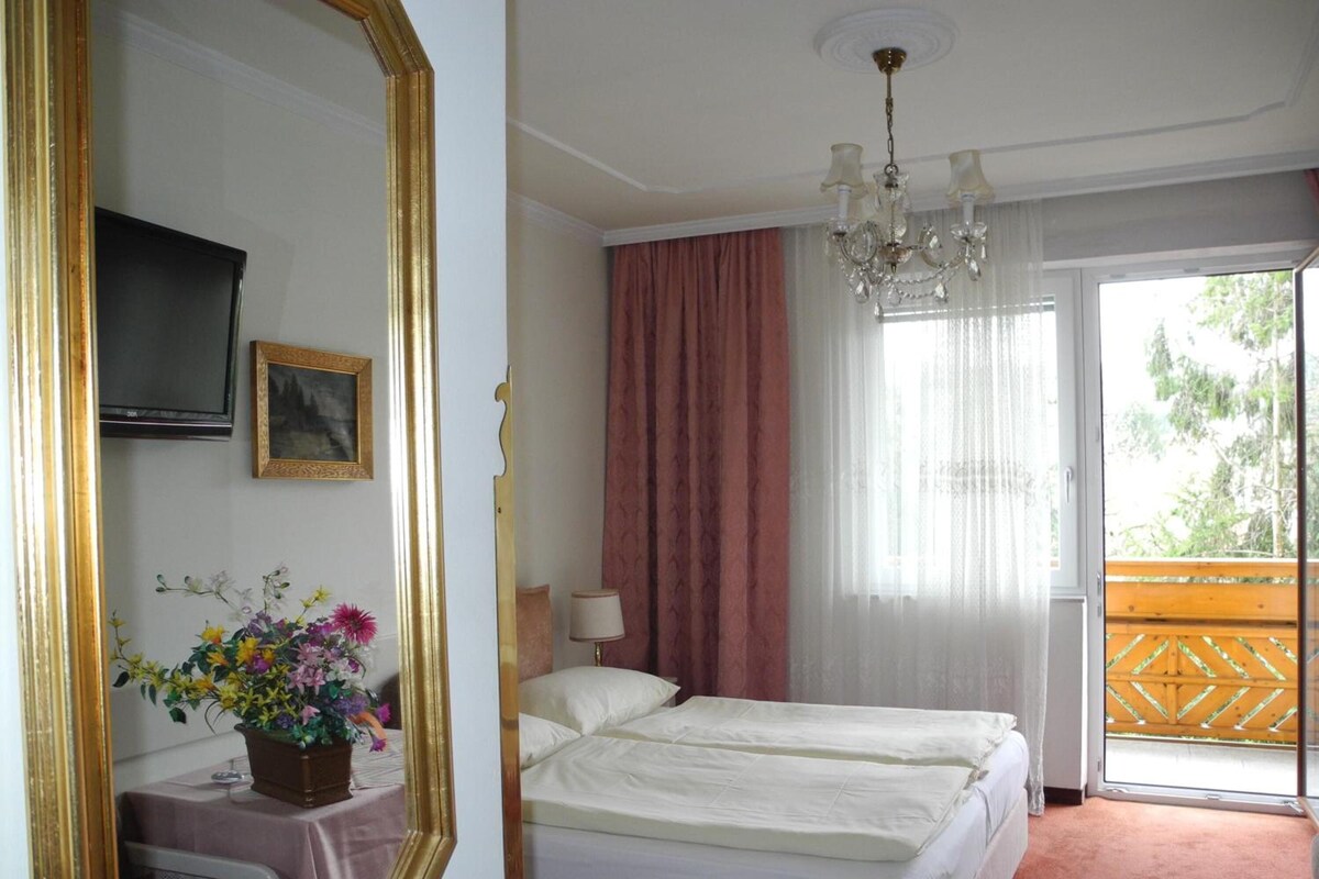 A hotel room with a bed, a television, a mirror, a window with curtains, and a balcony.