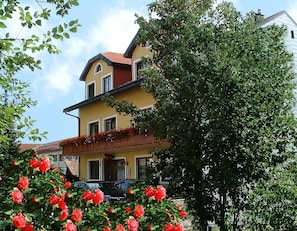 A two-story building with a balcony, surrounded by greenery and red flowers.