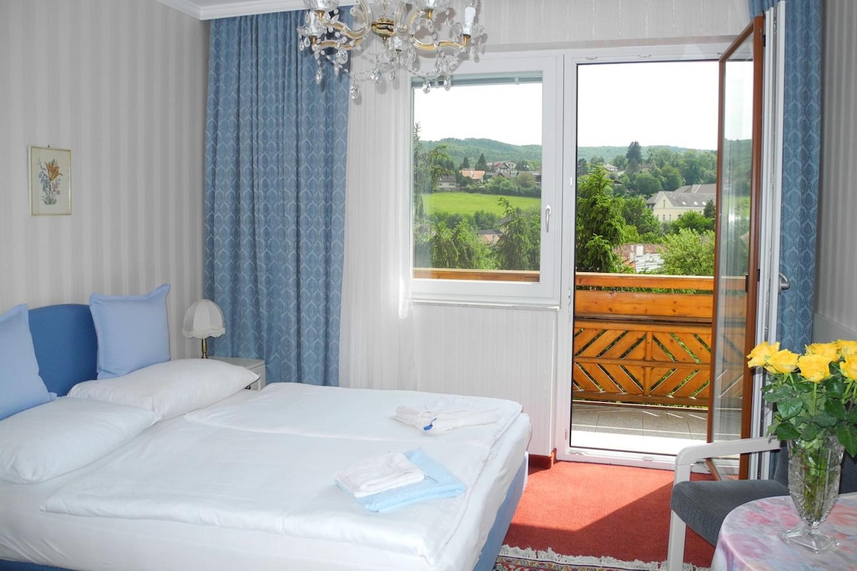 A hotel room with a bed, a chandelier, a view of greenery, and a balcony with a wooden railing.