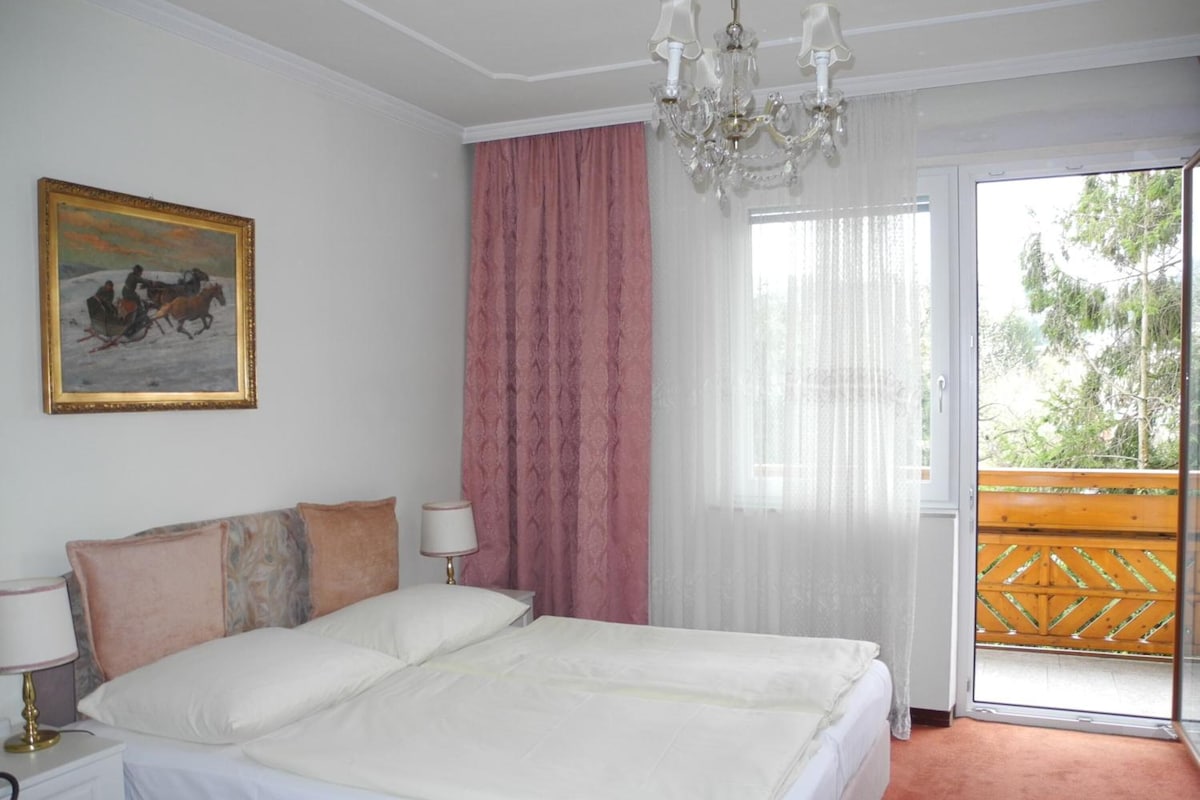 A bedroom with a bed, nightstands, a chandelier, a painting, and a view through a window.