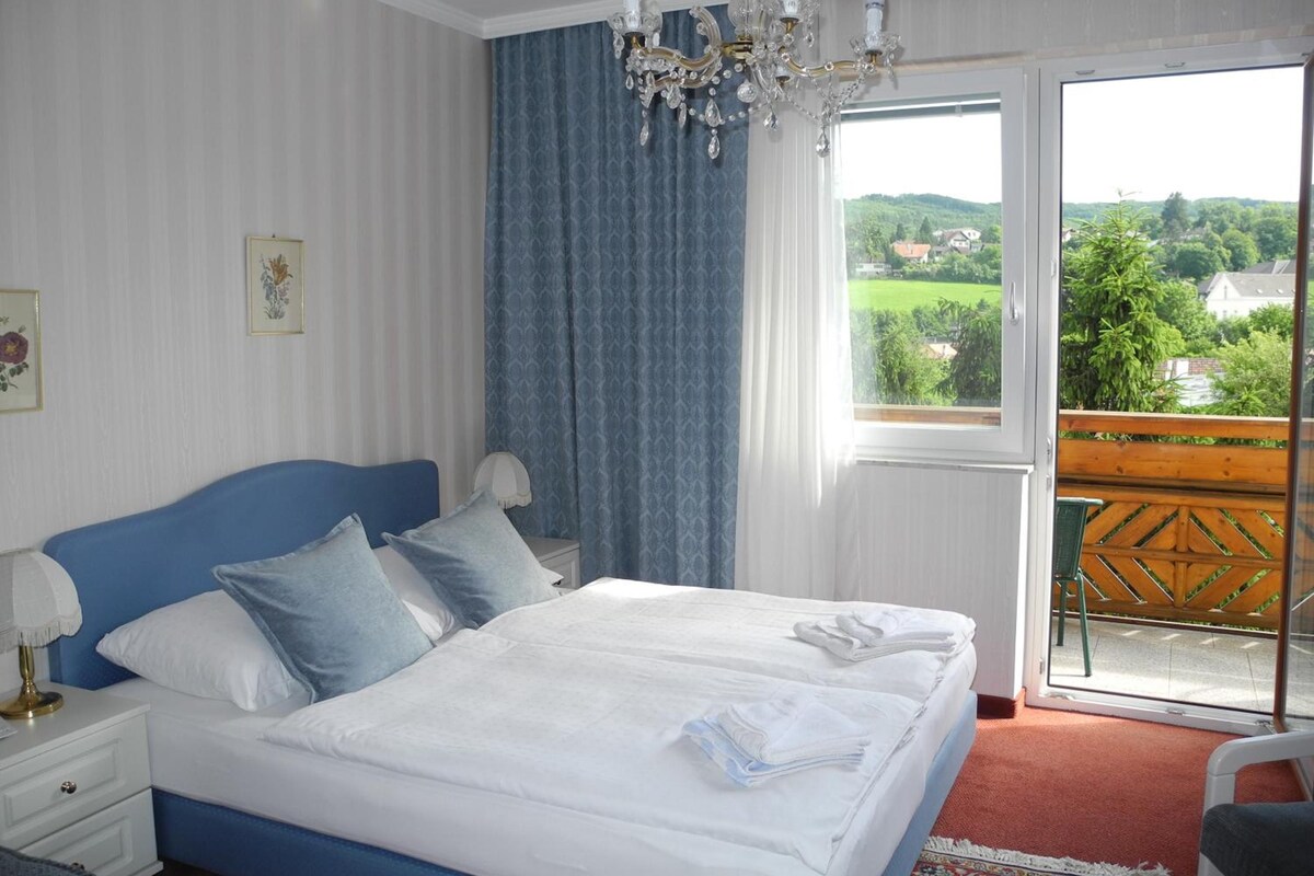 A hotel room with a large bed, a nightstand, a chair, a balcony with a view of greenery and houses.