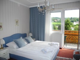 Habitación de hotel con una cama grande, una mesita de noche, una silla y un balcón con vistas a zonas verdes y casas.