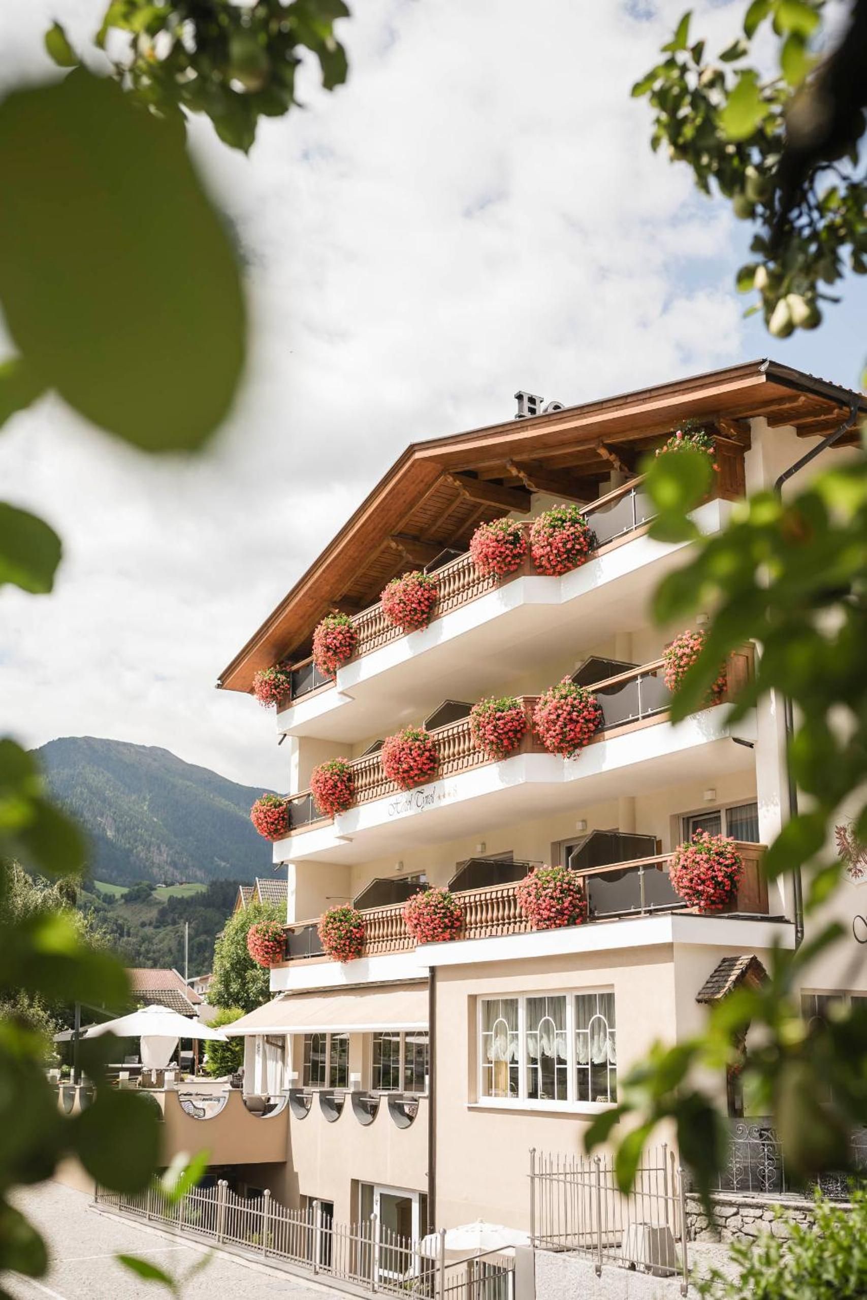 Un hotel a più piani con balconi decorati con cesti di fiori, sullo sfondo di montagne.