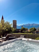 A hot tub with a wooden fence, lounge chairs, and a view of a castle and mountains.