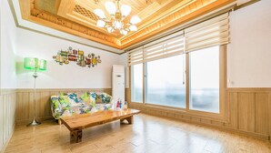 Une pièce spacieuse avec un plancher en bois, un banc en bois, un canapé coloré, un lampadaire vert et une grande fenêtre avec des stores.