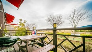 Une terrasse en bois avec des tables de pique-nique et un barbecue, donnant sur une zone herbeuse bordée d’arbres et sur une chaîne de montagnes lointaine.