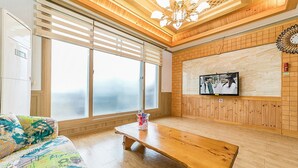 Un salon avec un banc en bois, un canapé coloré, un téléviseur fixé au mur et de grandes fenêtres équipées de stores.