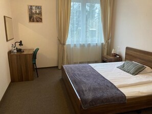 Quarto de hotel com cama, escrivaninha, cadeira, mesa de cabeceira, abajur, janela com cortinas e um quadro na parede.