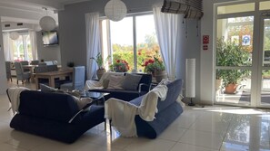 A modern living room with a blue sofa set, a dining area with a table and chairs, and large windows with curtains. - Hotel Solec (Gora Kalwaria)