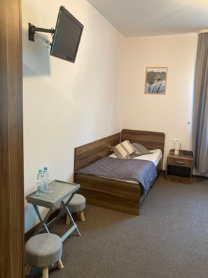 Quarto com cama, escrivaninha com cadeira, televisão fixada na parede e uma mesinha de cabeceira.