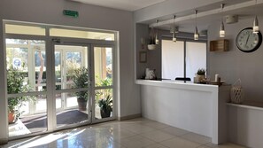 A reception area with a glass door entrance, a reception desk, hanging pendant lights, a wall clock, and potted plants.