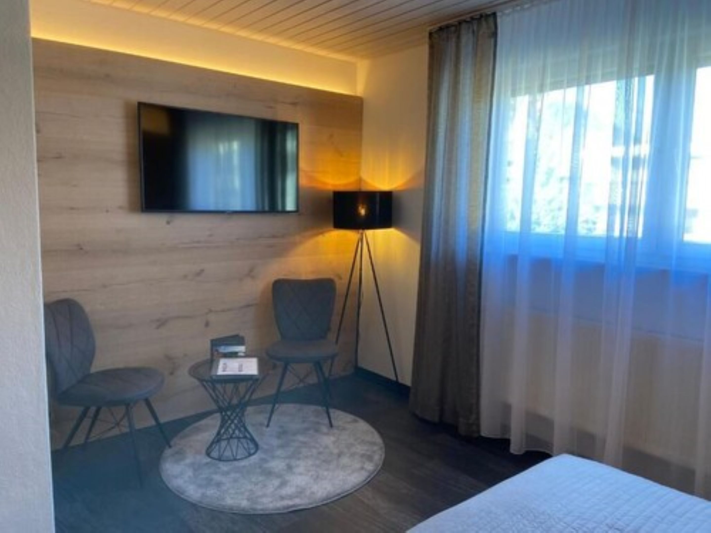 Habitación de hotel con una cama, un televisor montado en la pared, una lámpara de pie, dos sillas, una mesita y un ventanal con cortinas.