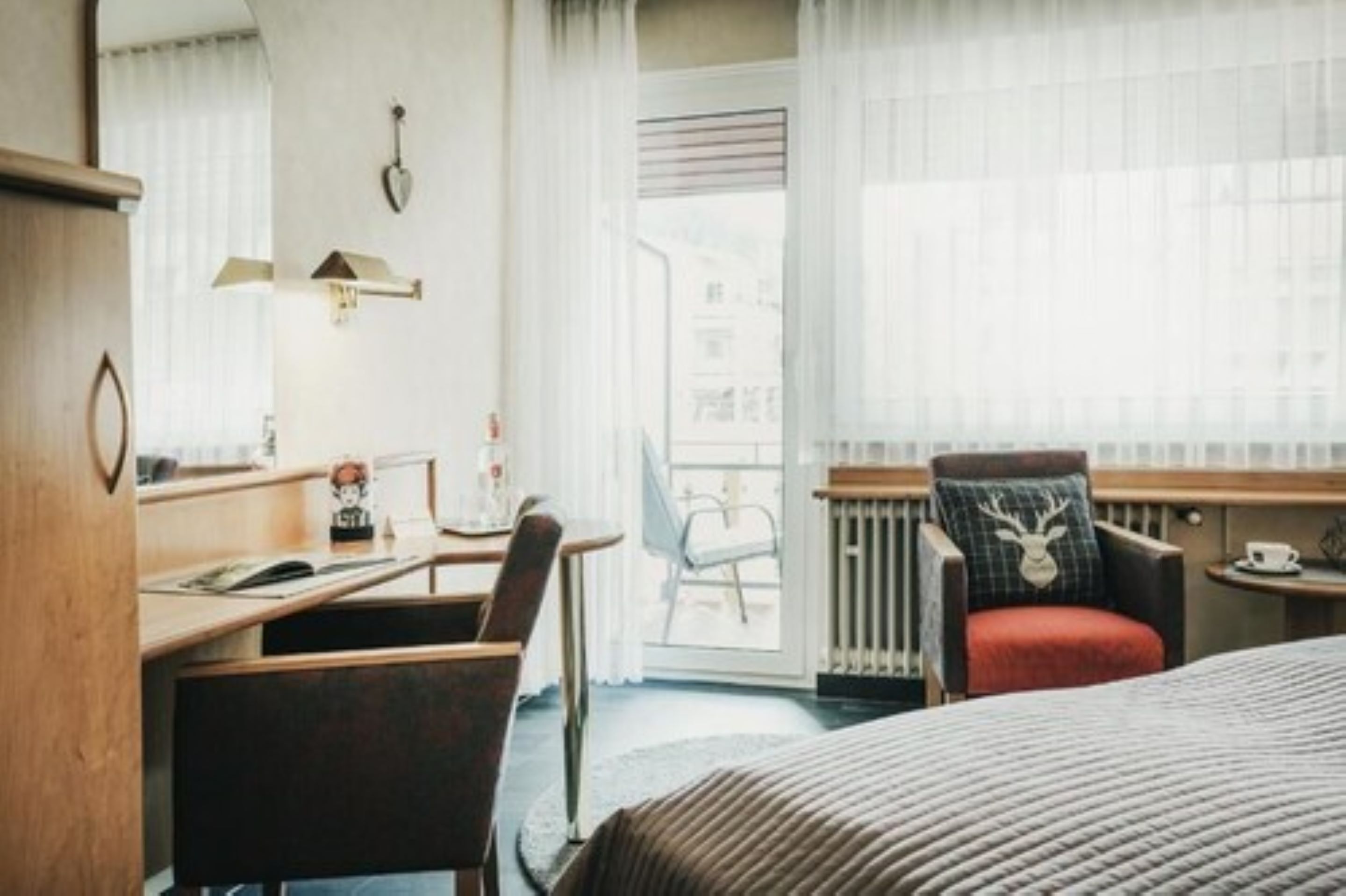 A hotel room with a bed, a desk, a chair, a radiator, and a window with curtains.