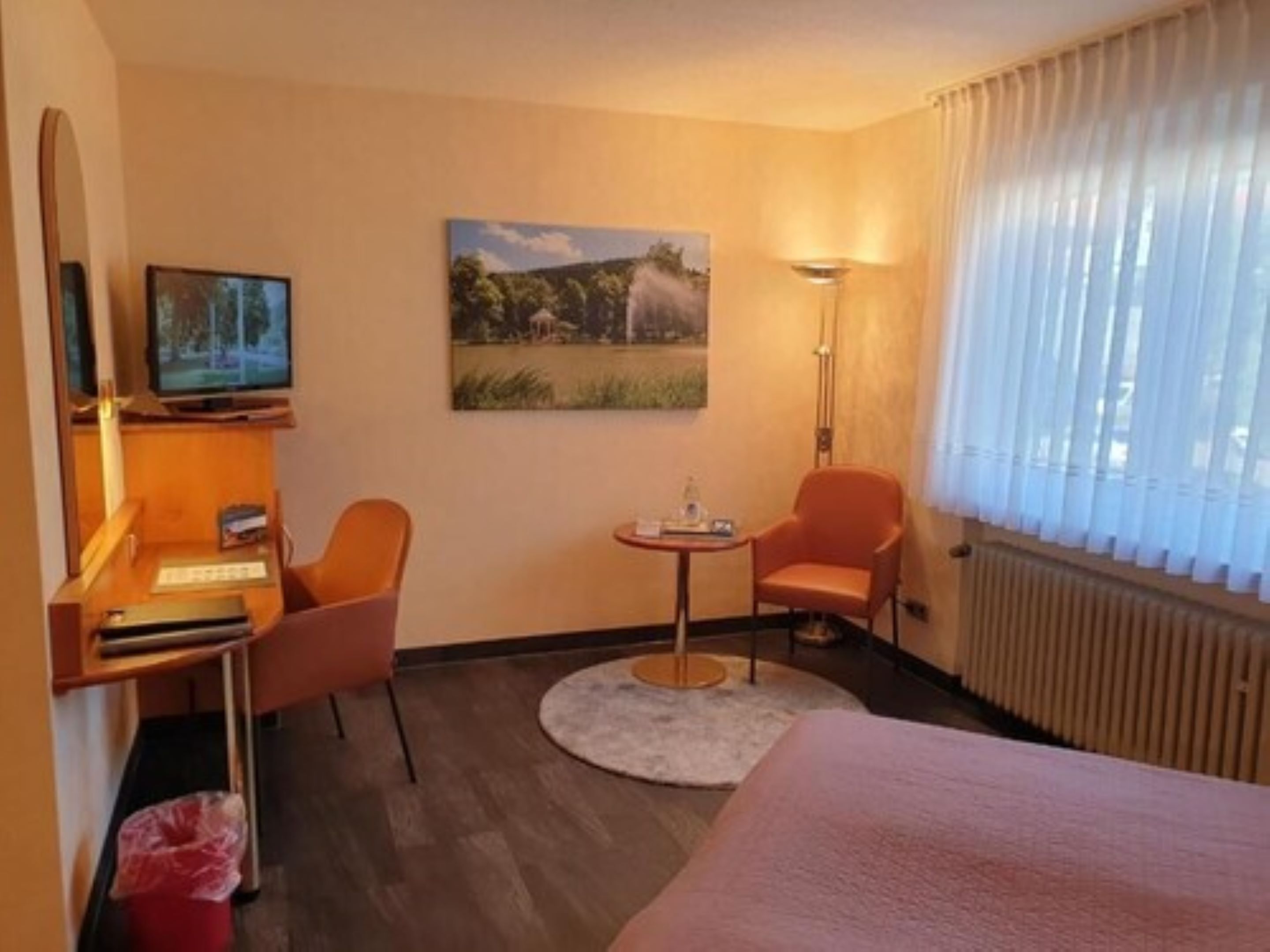 A hotel room with a bed, a desk with a computer, a chair, a small table, a painting on the wall, and a window with curtains.