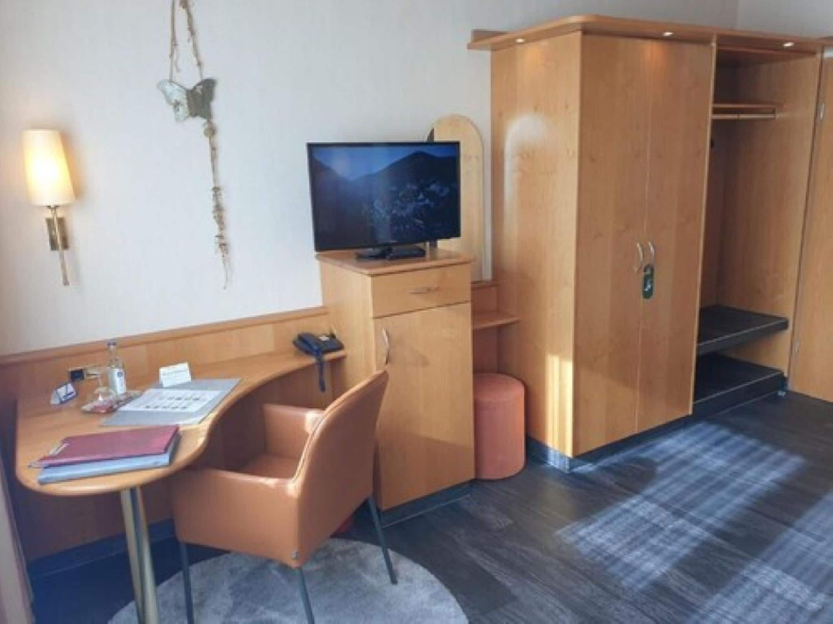 A compact hotel room with a wooden desk, a chair, a television, and a wardrobe.