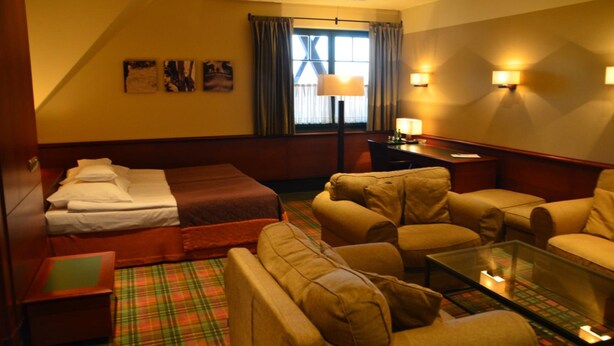 Una habitación de hotel con una cama, dos sillones, un sofá, una mesa de centro de vidrio y un escritorio con lámpara.