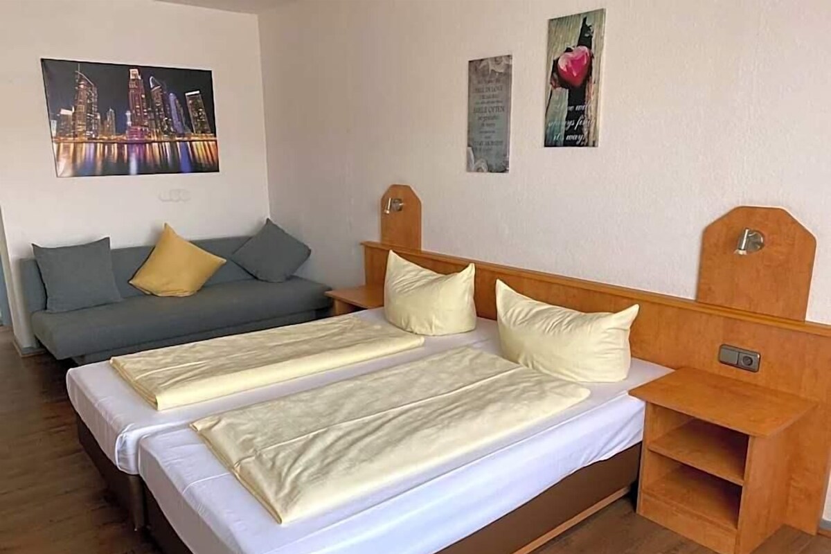 A hotel room with a large bed, a smaller sofa, a wooden nightstand, and a wall-mounted TV.