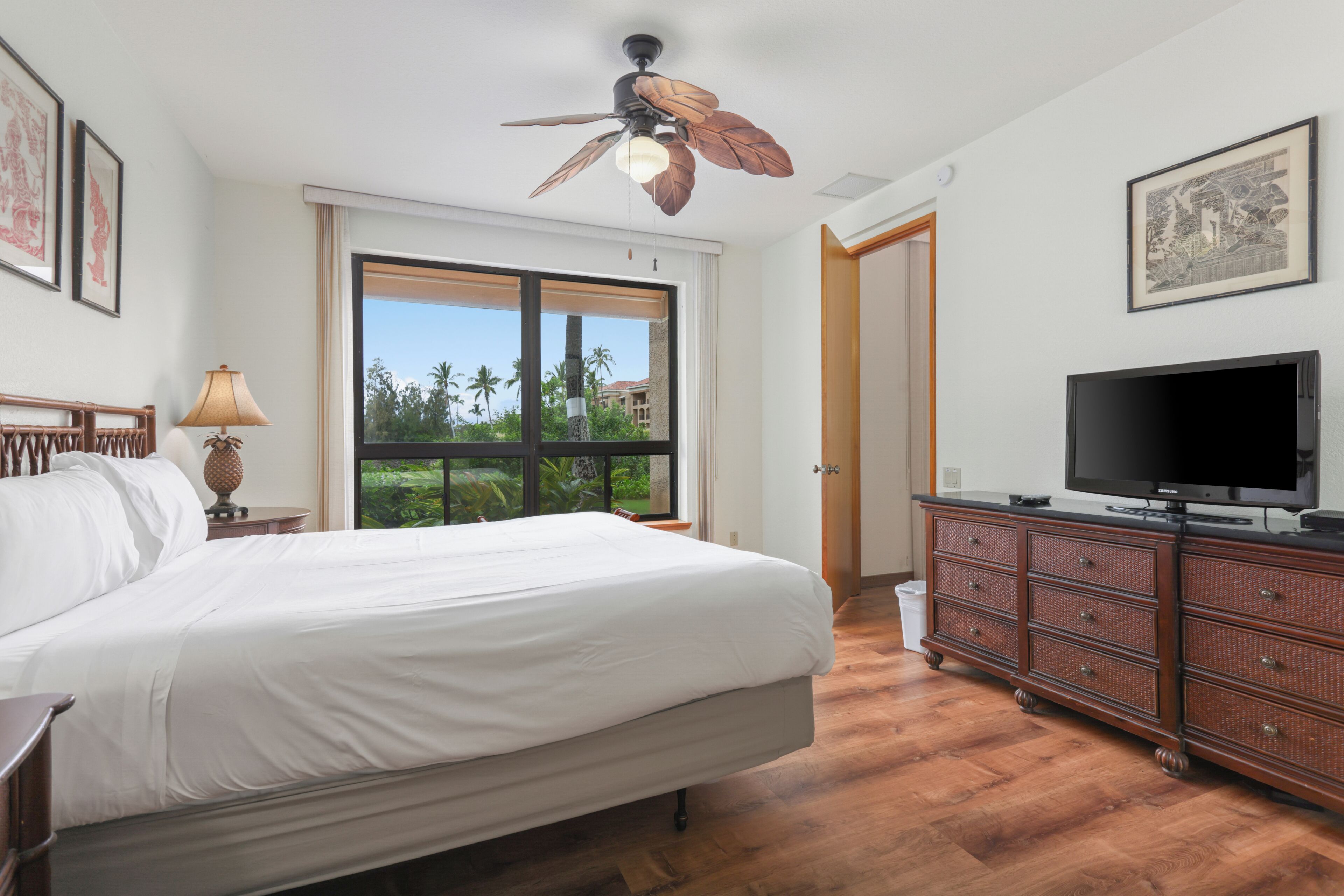 Une chambre à coucher comprenant un lit, une télévision, une commode, un ventilateur de plafond et une fenêtre donnant sur des palmiers.