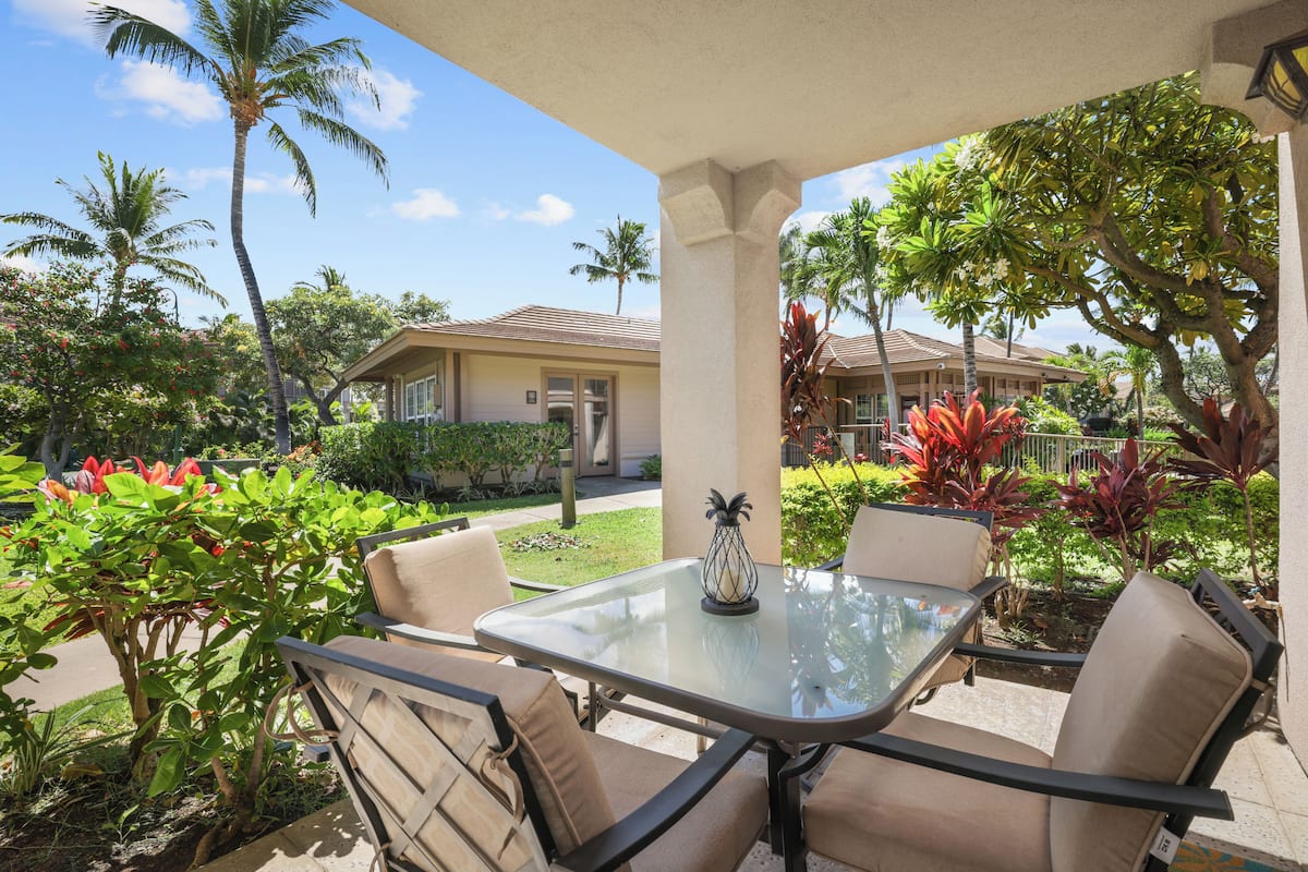 An outdoor patio with a glass table and two chairs, surrounded by tropical plants and palm trees.