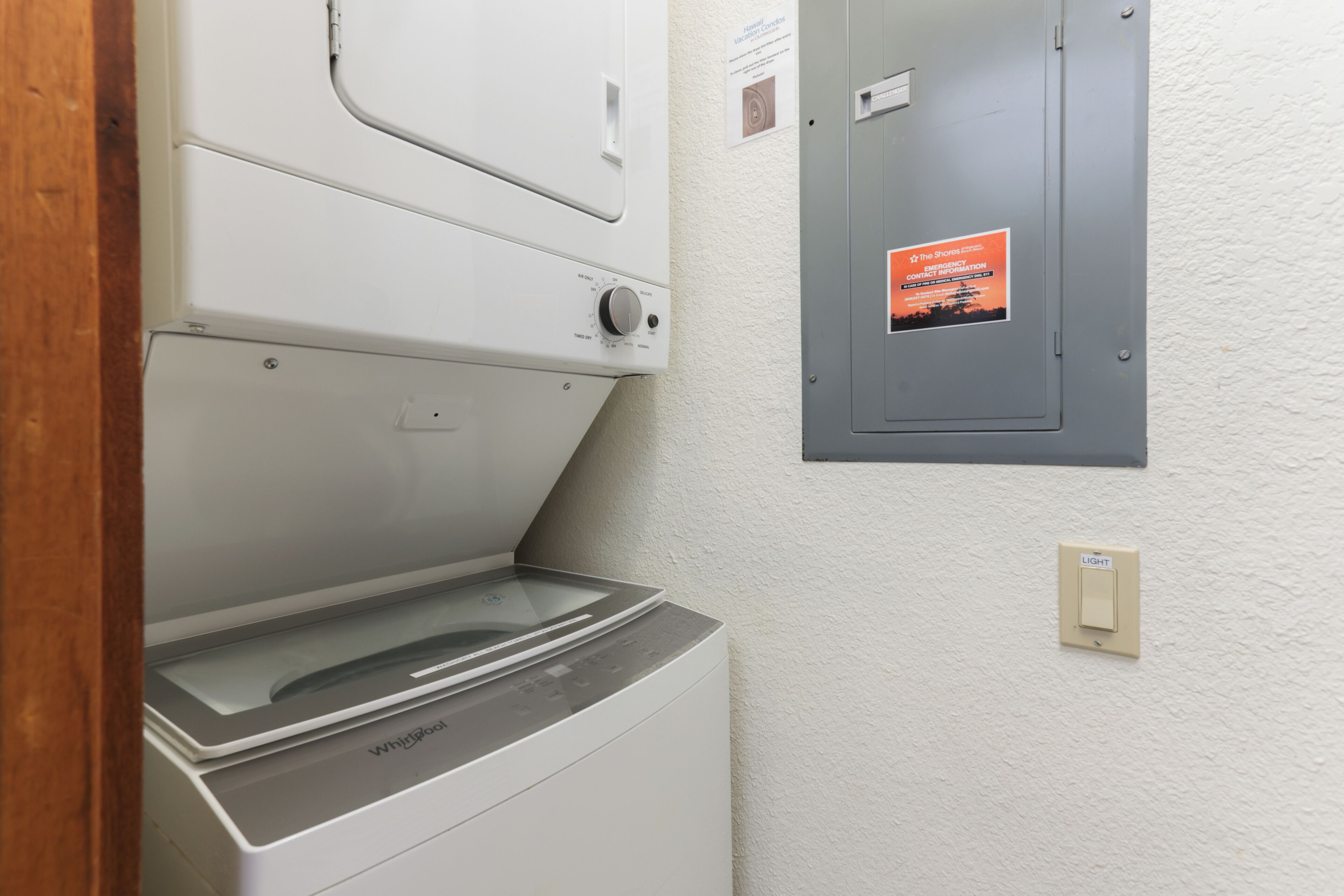 Un lavadero con lavadora y secadora Whirlpool, un interruptor de luz y un panel de servicios con un cartel de información de contacto.