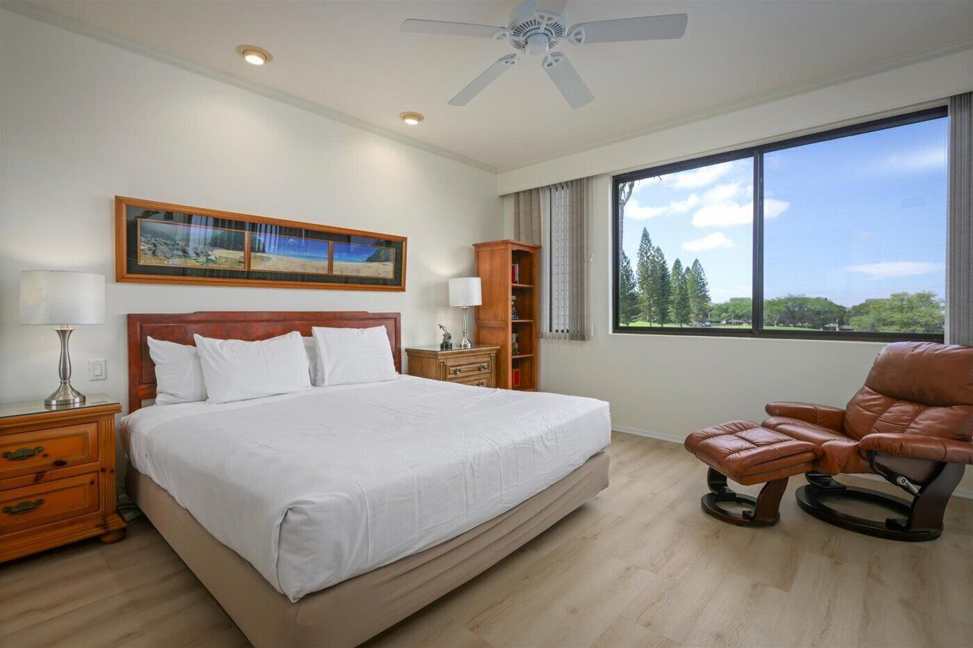 A hotel room with a large bed, a wooden headboard, a nightstand, a lamp, a bookshelf, a massage chair, and a large window with a view of trees.