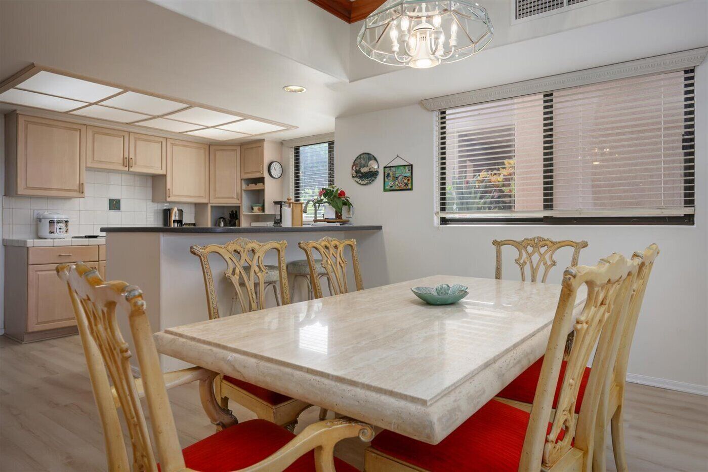 A modern kitchen with light wood cabinets, a central island, and a dining area with a marble table and red cushioned chairs.
