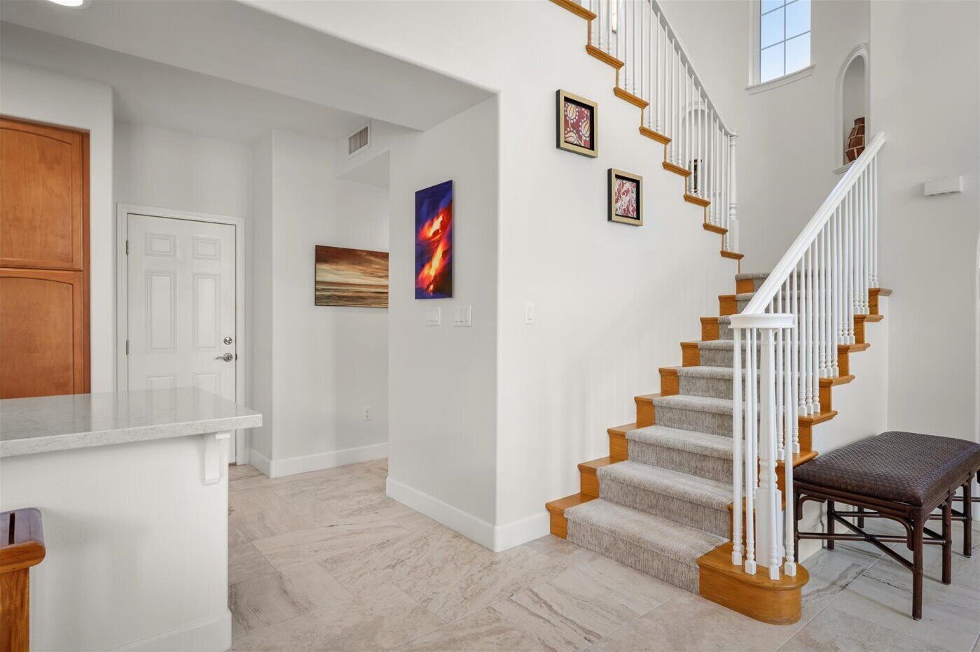 A spacious hallway with a staircase leading to an upper floor, a wooden bench, and framed artwork on the walls.