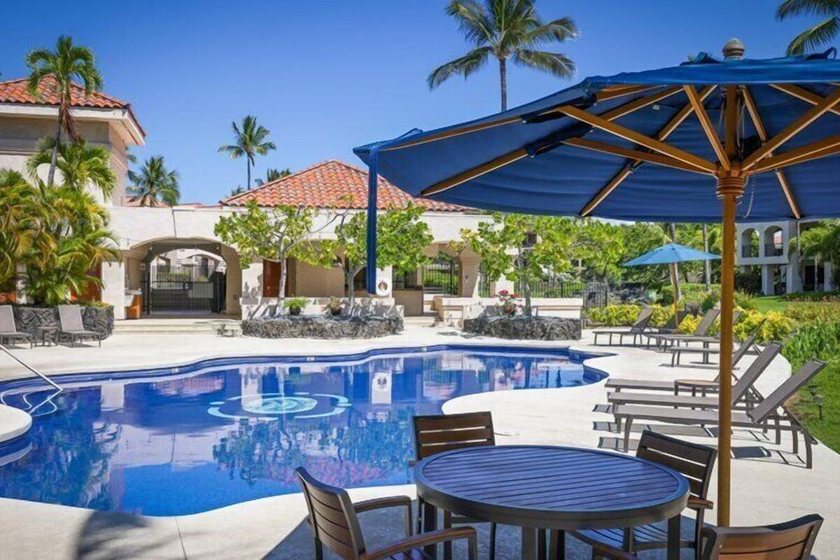 A swimming pool with lounge chairs and umbrellas, surrounded by palm trees and a building with a tiled roof.