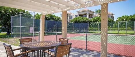 A covered outdoor seating area with a tennis court in the background.