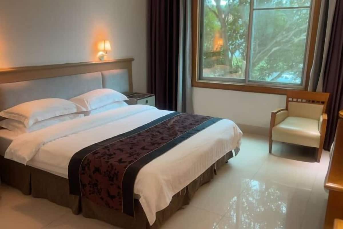 A hotel room with a bed, a nightstand, a chair, and a window with a view of trees.