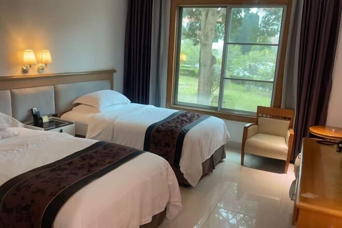 A hotel room with two beds, a chair, a small table, and a window with a view of greenery.