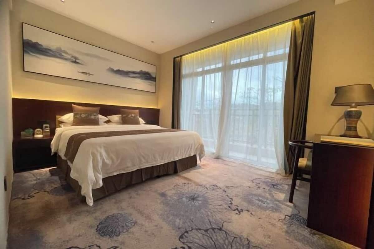 A hotel room with a large bed, a nightstand, a desk, a lamp, and a large window with curtains.