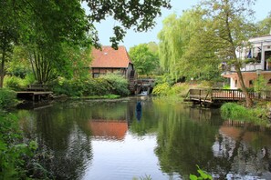 Un entorno tranquilo junto al río con un puente de madera, una casa de techo rojo y abundante vegetación.
