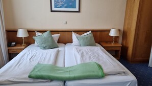A hotel room with two beds, each with white linens, green pillows, and a green towel. There are bedside lamps, a phone on the left nightstand, and a framed picture on the wall. - Residenzia Hotel Grenadier (Munster im Heidekreis)