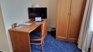 A small room with a wooden desk, a television, a chair, and a wardrobe. - Residenzia Hotel Grenadier (Munster im Heidekreis)