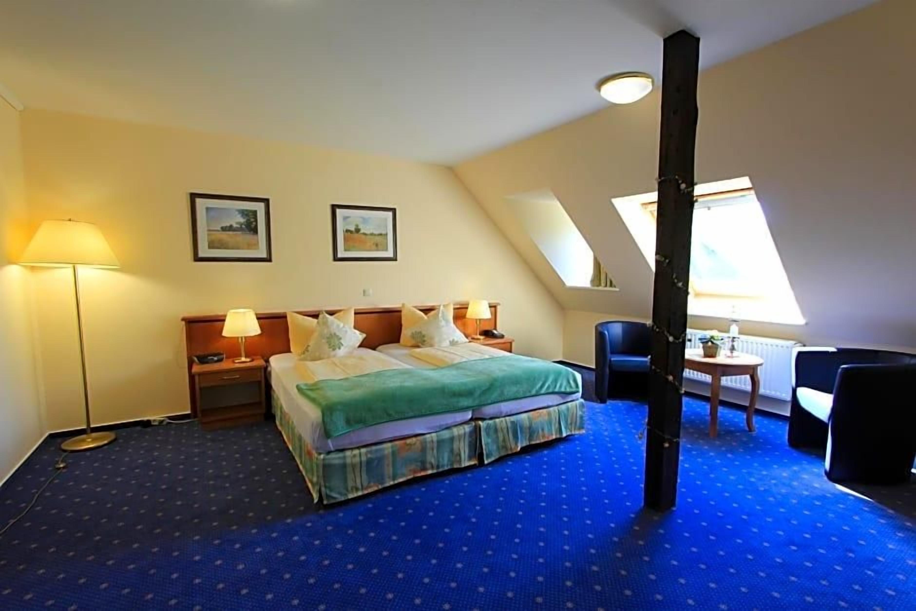 A hotel room with a bed, two bedside tables, a desk with a chair, a lamp, and two framed pictures on the wall.