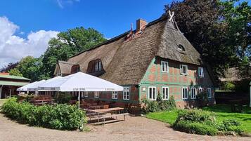 Ein traditionelles Gebäude mit einem Reetdach, einer grün-roten Fassade und Außenplätzen mit Sonnenschirmen.